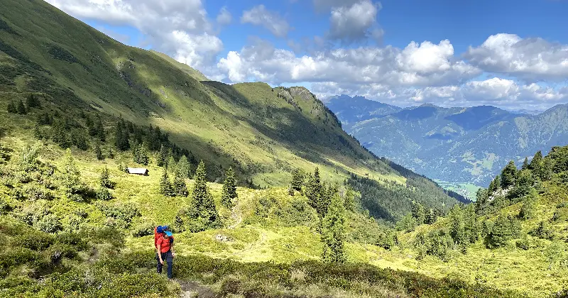 Een wandelaar in een rode jas loopt door een weelderige, groene bergvallei met hier en daar wat bomen en bergtoppen in de verte, onder een gedeeltelijk bewolkte hemel.